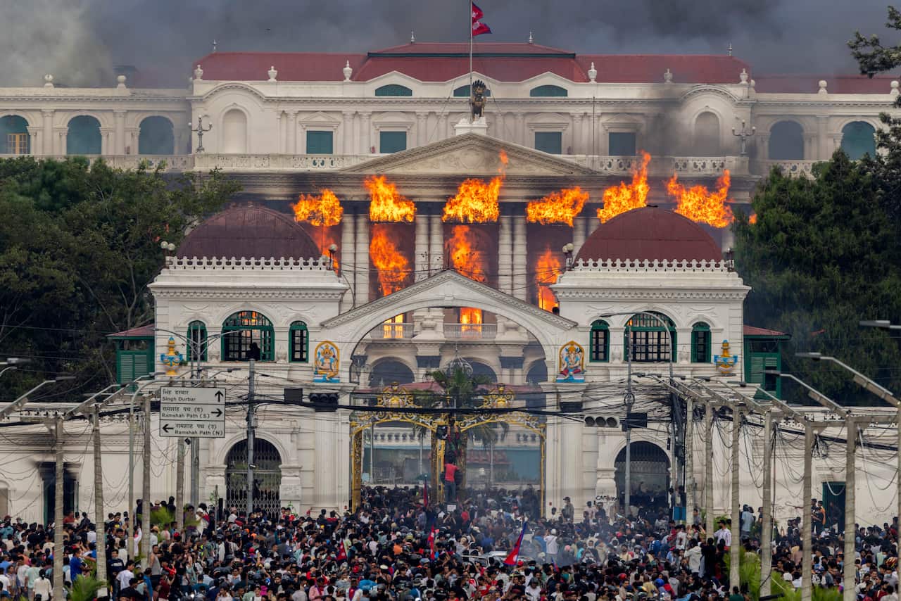A large crowd is standing in front of a white palace-like structure, partly on fire.