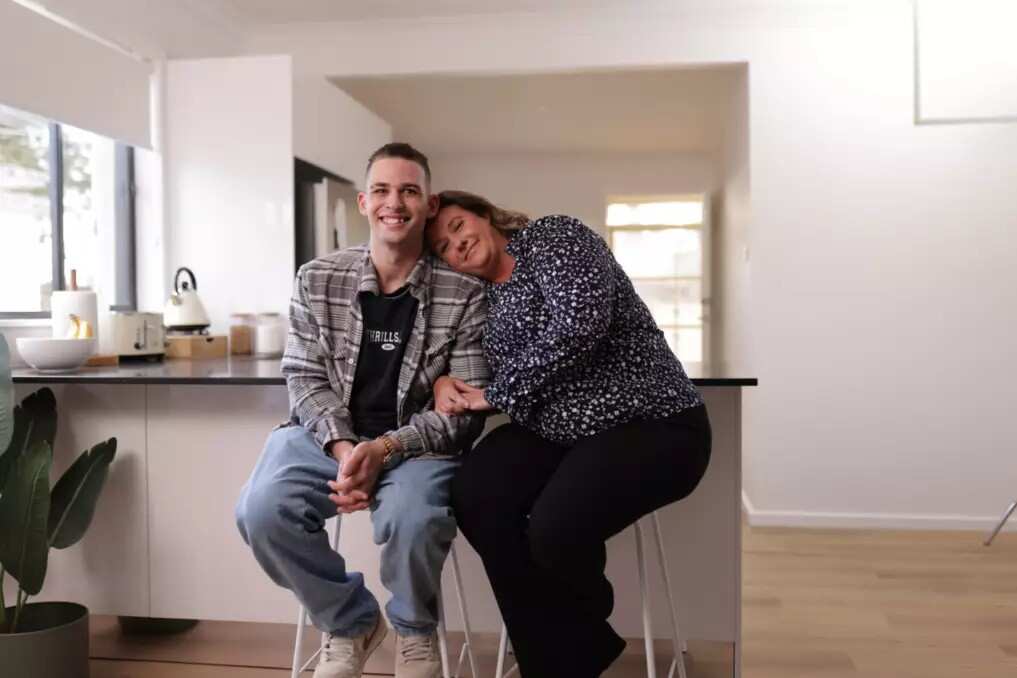 A woman is sitting with her head resting on her son's shoulder in a living room.