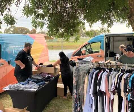 Two vans with a river in the background. Two people are standing at a table covered in clothes and there is a rack of clothes in the foreground.