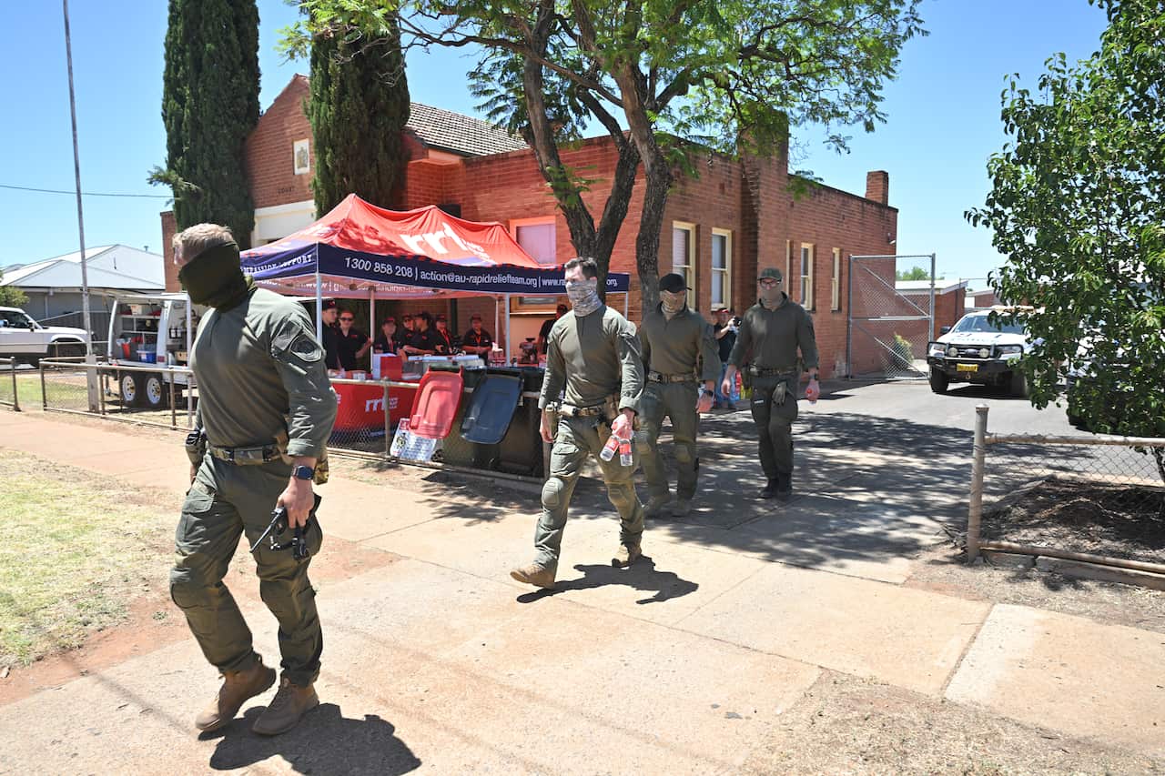 Four men in olive-green tactical gear and face gaiters walk along a paved path in front of a red brick building and a Rapid Relief Team tent.