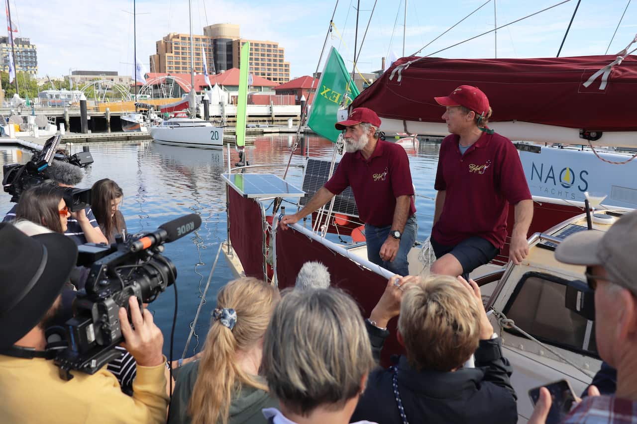 Two men wearing matching maroon shirts and caps stand on the edge of a yacht talking to the media