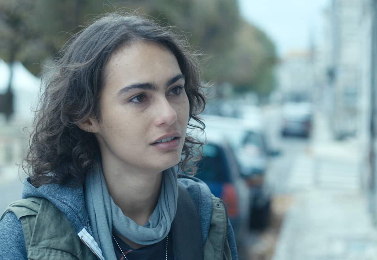 Woman with tousled hair wearing jacket and scarf looks to the right with a serious expression. 