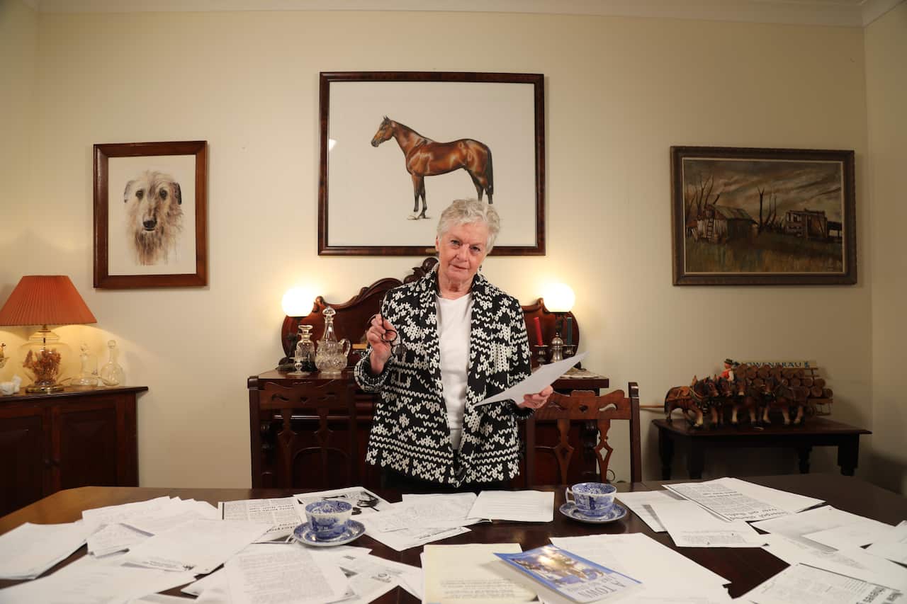 A woman stands behind a large desk covered in papers