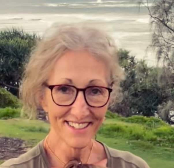 An older woman with glasses and wavy, grey hair tied in a bun stands in front of a row of trees beside the ocean