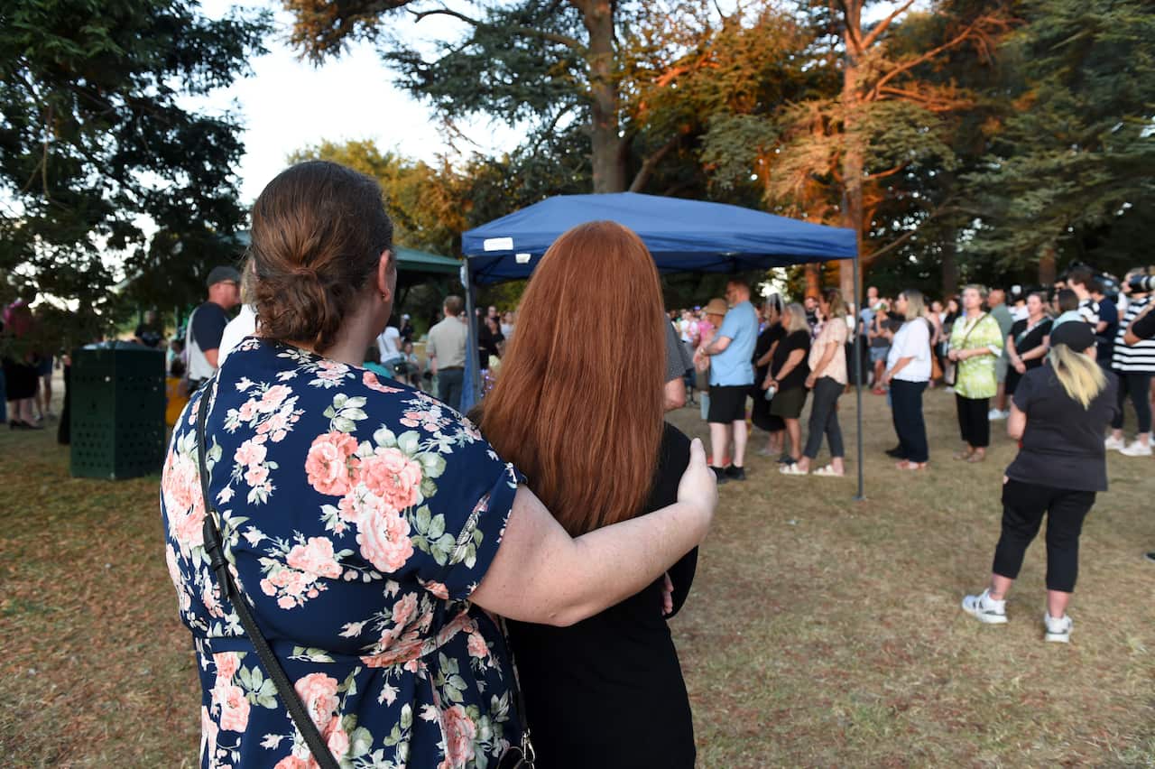 A woman in a floral dress embraces a woman in black