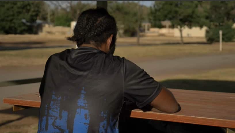 A photo of a man wearing a dark t-shirt and seated at a picnic table, taken from behind.