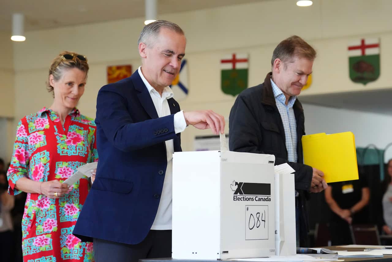 A man with white hair in a blue suit drops a piece of paper into a ballot box. There is a man carrying a yellow box next to him, and a woman in a red, print-patterned dress is standing behind him.
