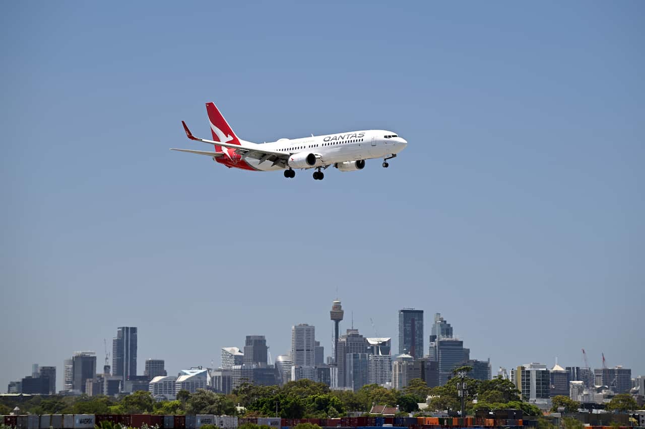 A white plane with a red tail flying with the city of Sydney in the background