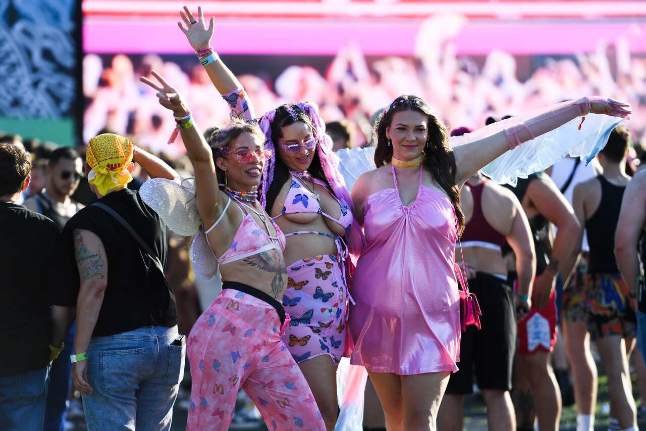 Three people in flamboyant pink party outfits with bitterfly motifs pose with arms outstretched at a concert.