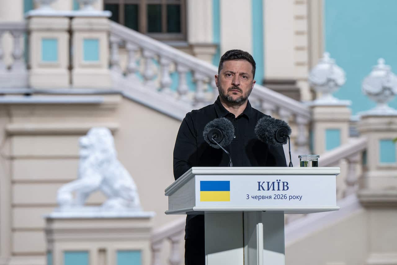 Volodymyr Zelenskyy, a middle aged man with black hair and a short beard, dressed in black, stands at a podium speaking into microphones.