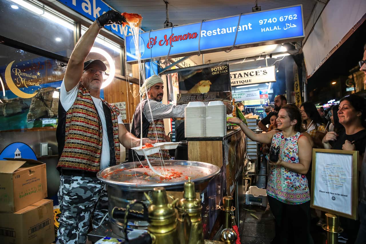 A food vendor putting orange sweets in a disposable box as customers wait. There's another vendor standing next to him in front of a counter.