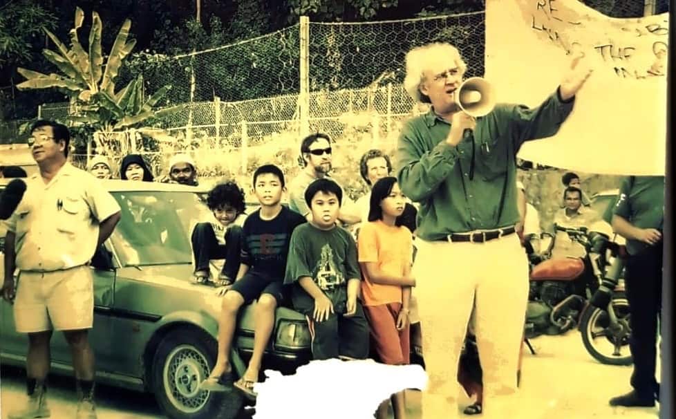 An old photo of an old man is speaking into a handheld microphone cum speaker with one hand extended in the air. Children sit on a car's bonnet behind him, along with some adult men.