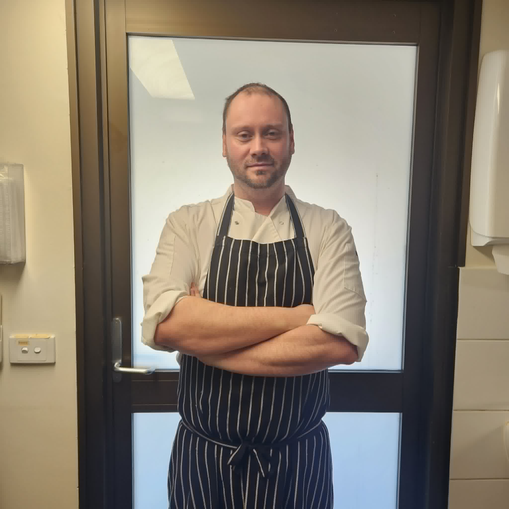 A man wearing a chef's uniform stands in front of a door with his arms crossed