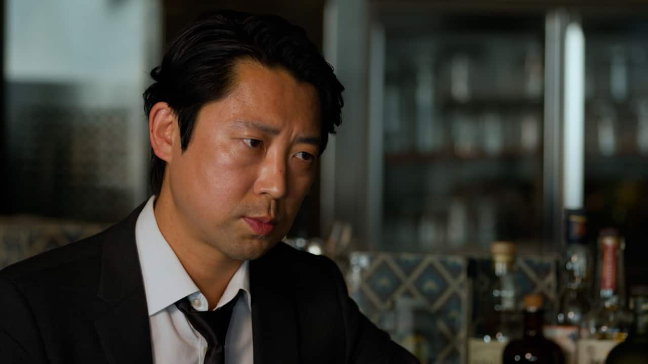 A man with short dark hair and a wearing a suit and black tie looks at the camera with a serious expression.