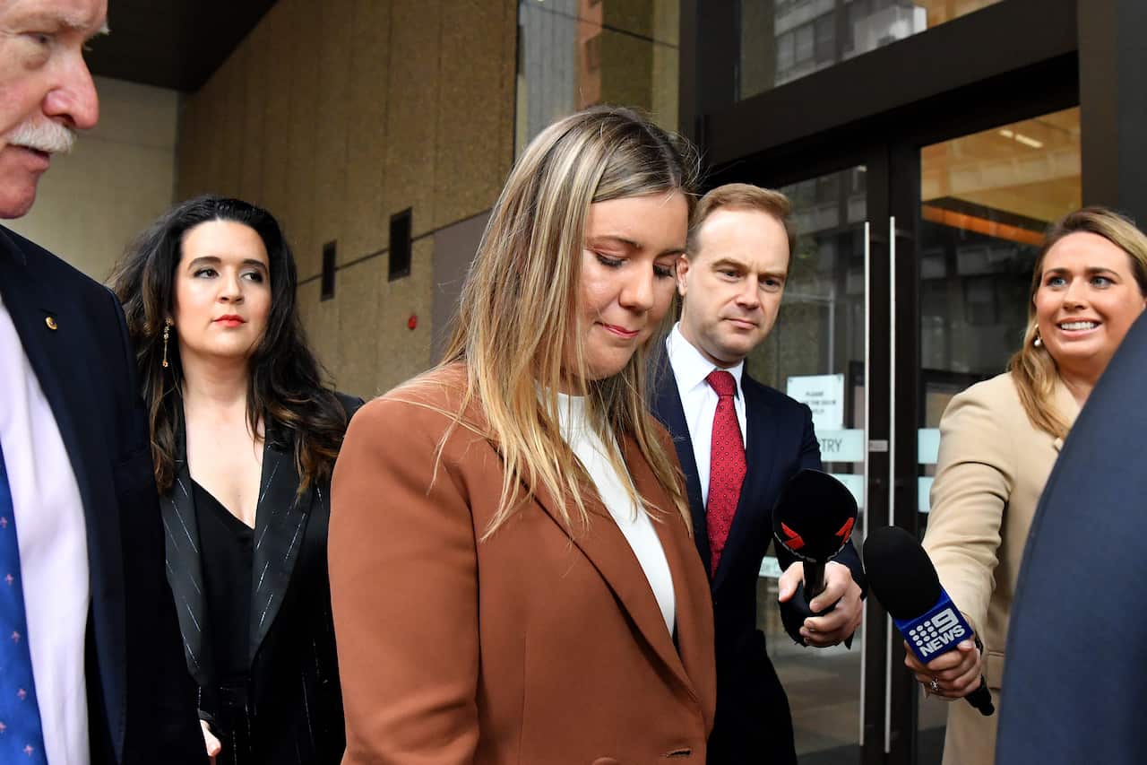 A woman with her head down walks as journalists hold microphones in front of her.