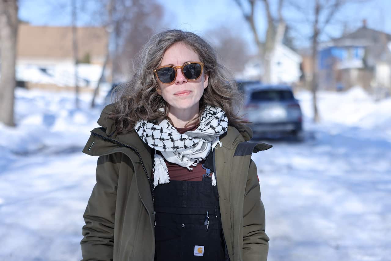 A blonde woman wearing sunglasses, a keffiyah and a thick coat stands in the middle of a snow covered suburban street. 