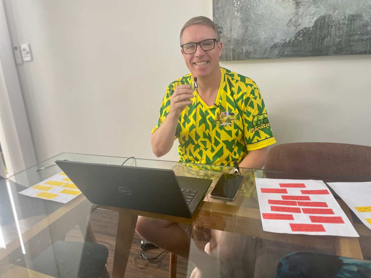 David Feeney wearing a green and yellow patterned football jersey while sitting behind a laptop at a glass-topped table. He is holding a small microphone to his mouth.