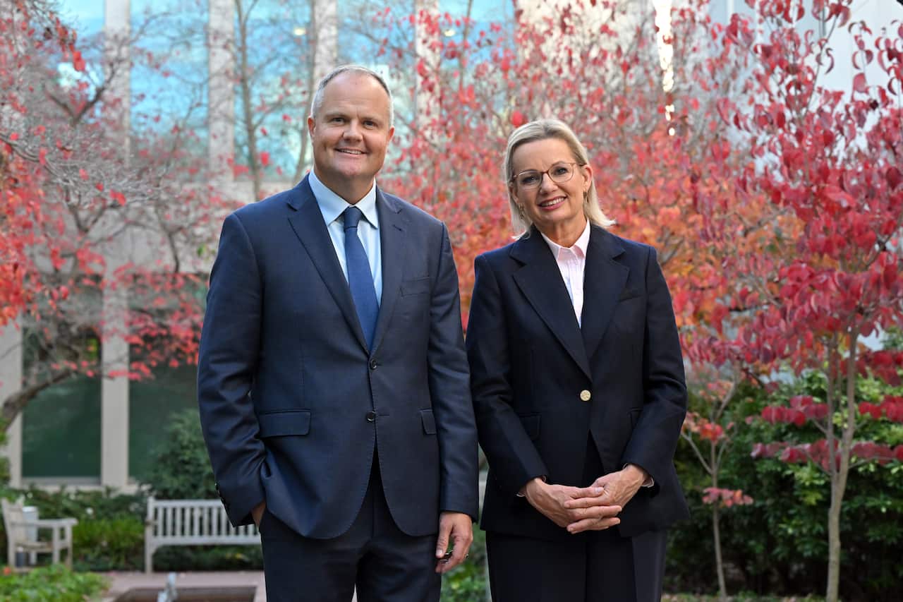 A man and a woman in suits stand in front of red autumn leaved trees.