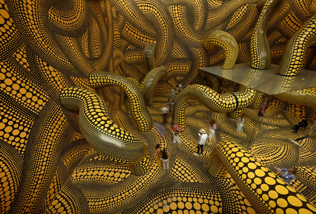 A tangle of black and yellow tendrils fill a room. People are walking on some of them.