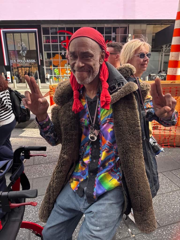 Man wearing a colourful patterned shirt, red headscarf and shearling-lined coat poses on a city street, holding up peace signs with both hands.