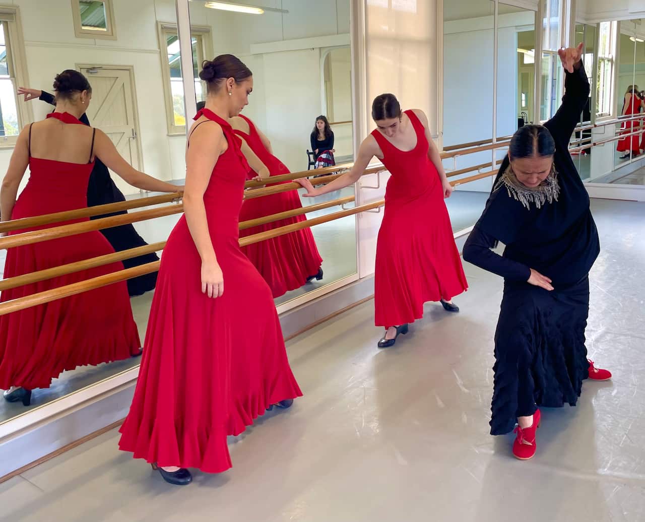 Three female dancers in a studio. Two are wearing red flamenco dresses and standing near a mirror. One is is standing in front of them and wearing a black flamenco dress.