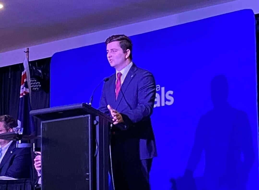 Young man in suit standing in front of a Liberal Party banner, making a speech.
