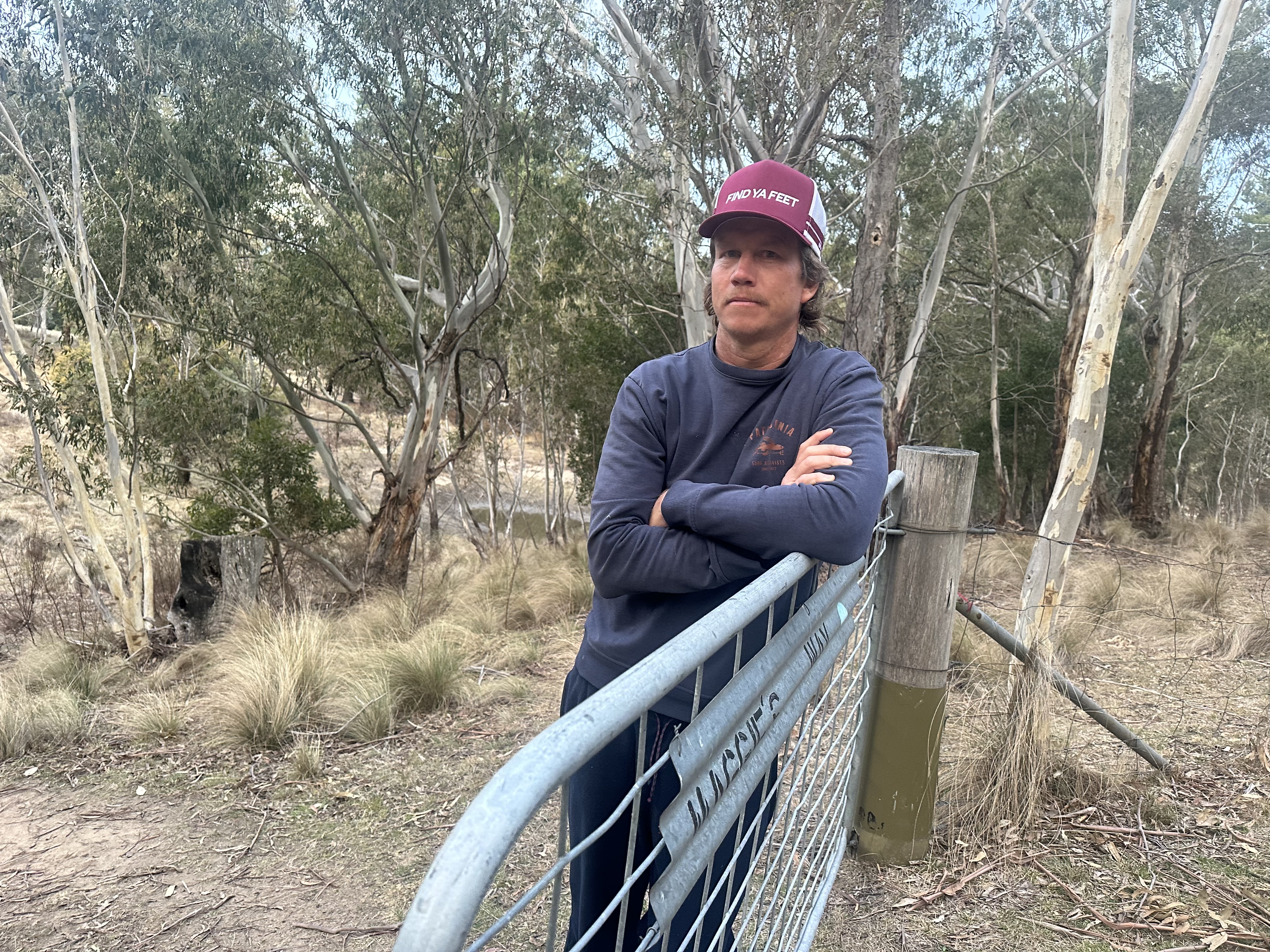A man in a cap leans on a gate in the country with a serious expression.