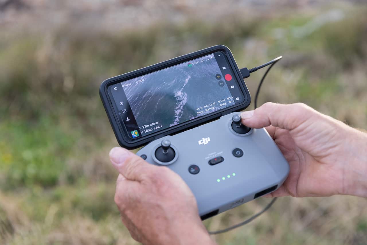 A close-up shot of a person's hands operating a DJI drone controller with a smartphone attached, displaying a live feed of waves from the drone's camera.