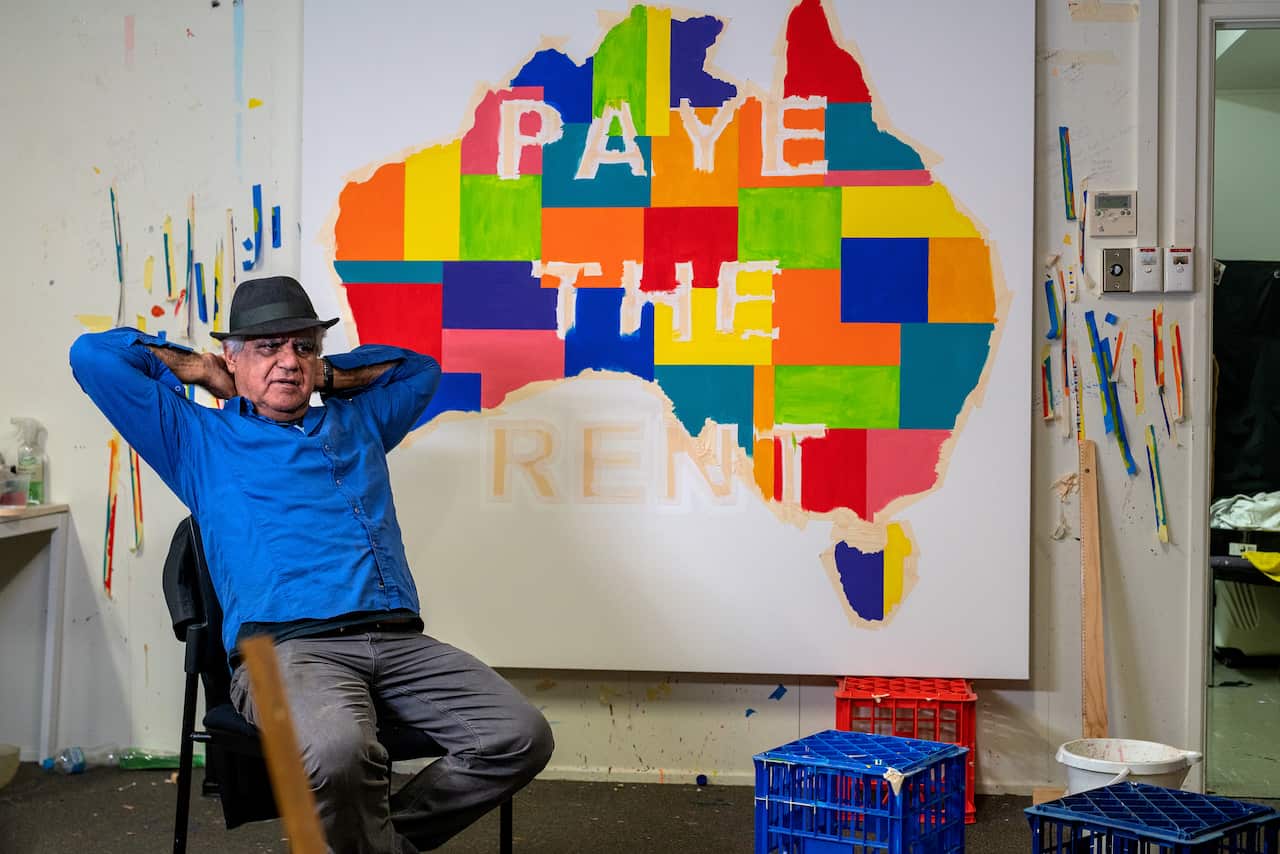 Richard bell reclines in a blue shirt and fedora in front of a painting, a multicoloured patchwork of australia with the words 'Pay the rent' superimposed on them. 
