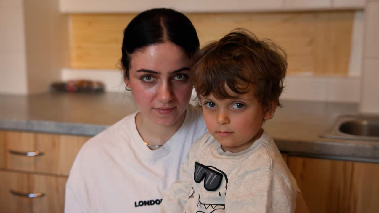 A young woman and a small child look directly at the camera with serious expressions while posing together in a kitchen.