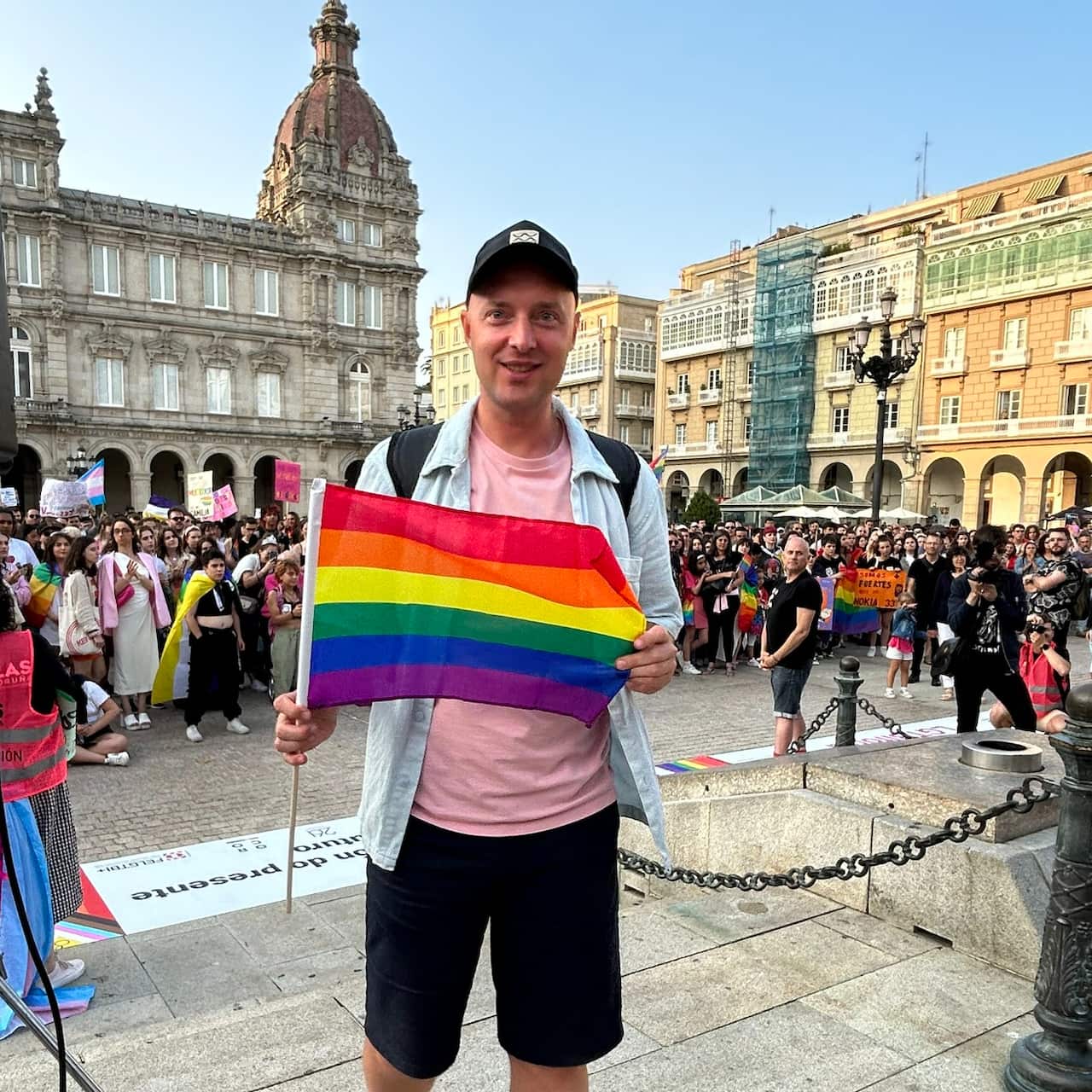 A man wearing a light blue shirt over a pink t-shirt, black shorts, a baseball cap and a backpack standing outside in a square while holding a small rainbow flag