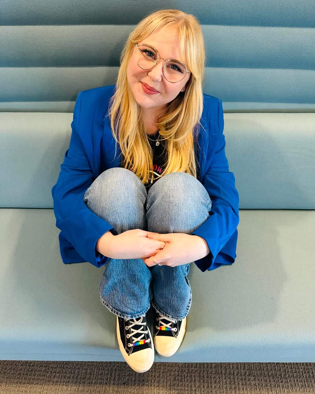 A young girl is wearing jeans and a blue shirt, sitting down on a couch smiling with her hands joined