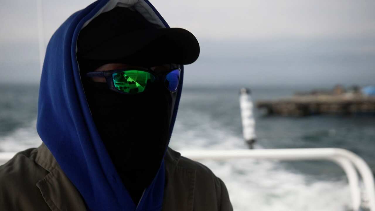 A man wearing a blue hoodie, a black balaclava, and sunglasses to conceal his face. 