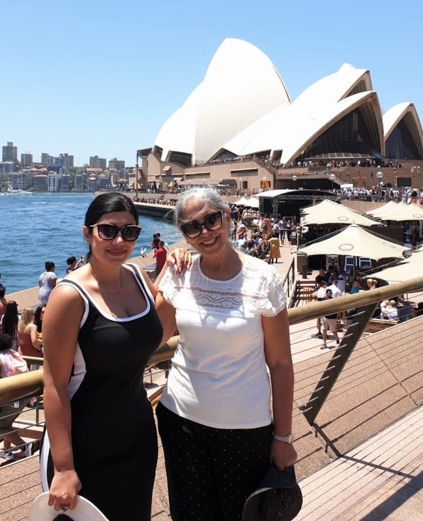 A woman in a black top standing with a woman in a white t-shirt in front of the Sydney Opera House. 