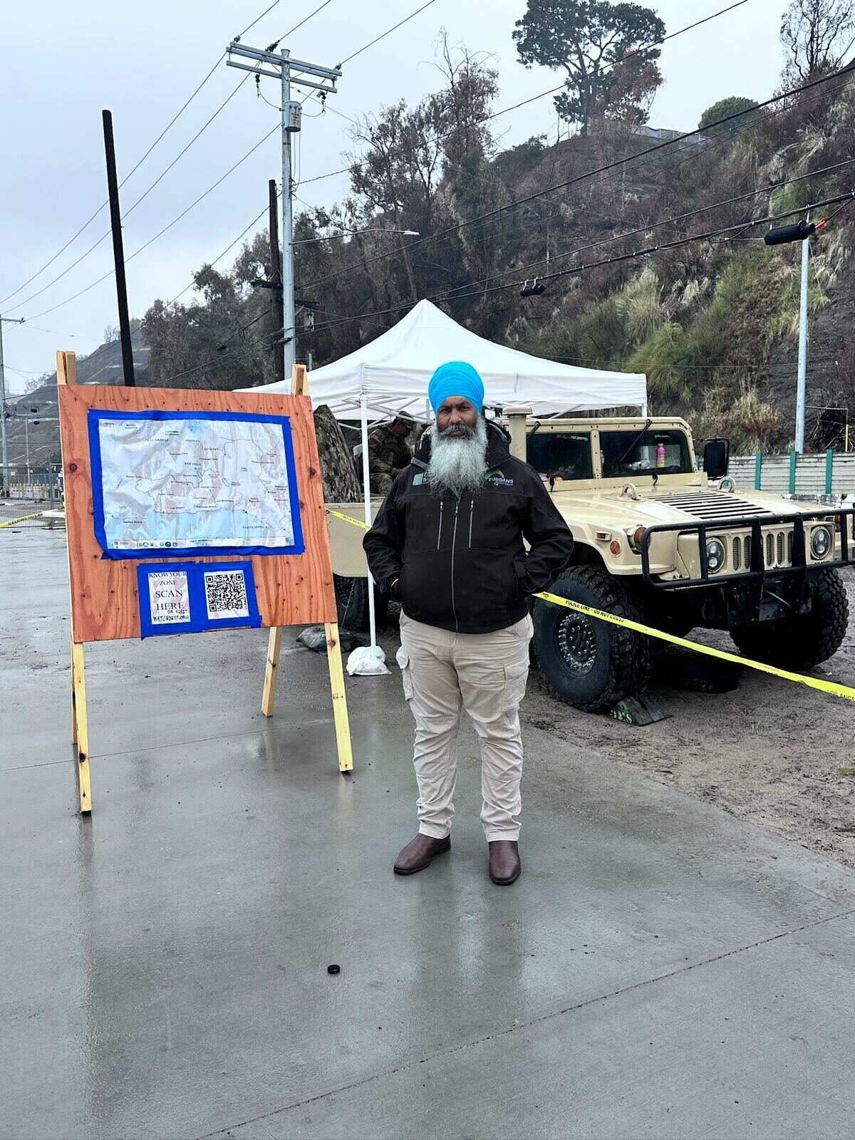 A Sikh man, wearing a blue turban and a long beard, stands outside with a truck behind him.