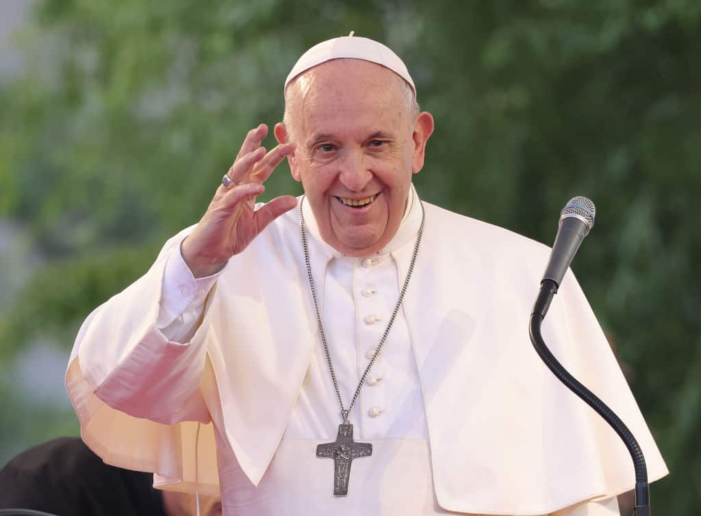 Pope Francis, wearing all white with a silver crucifix on a chain around his neck, waves and smiles as he stands behind a microphone