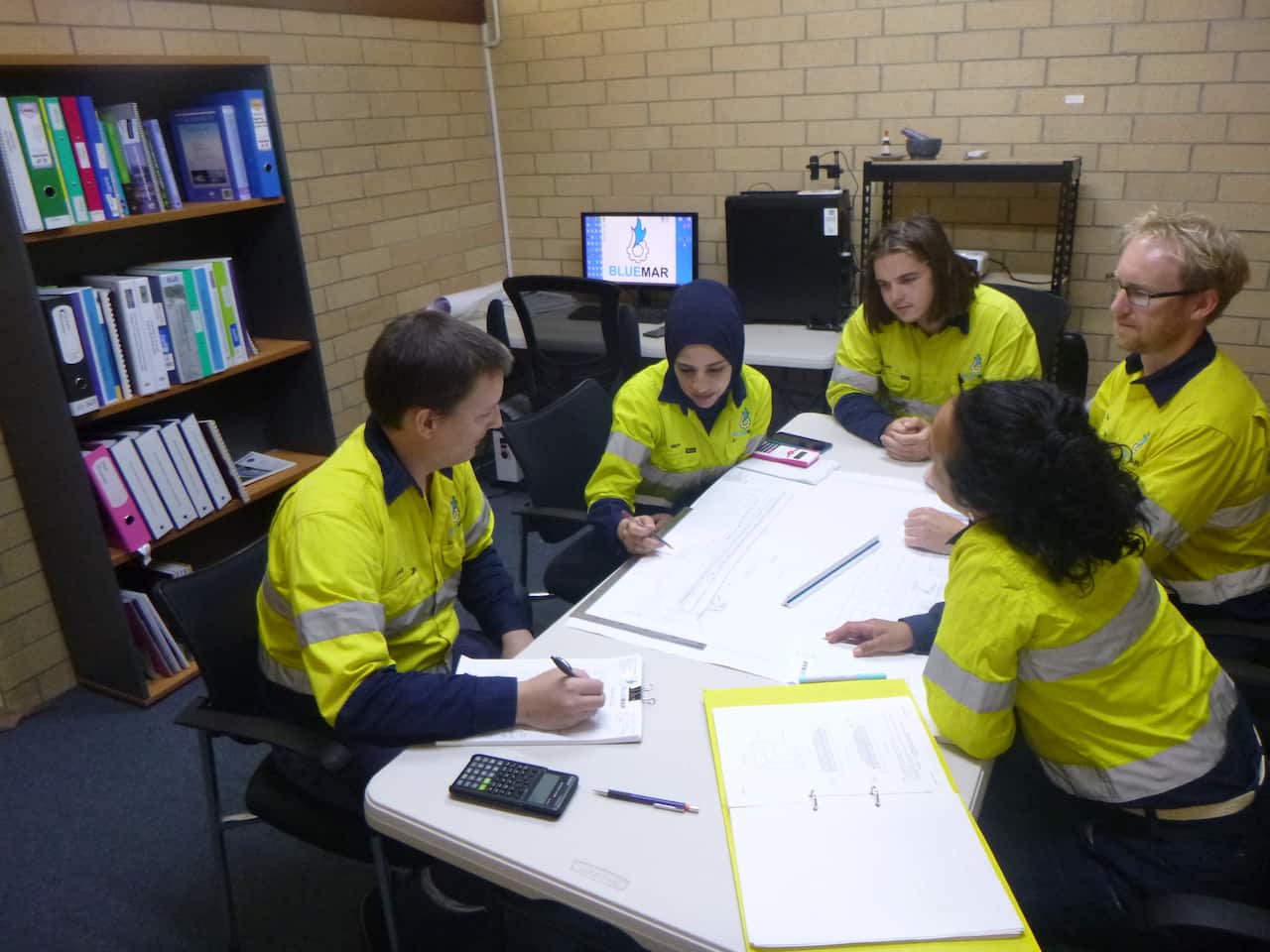A group of people in high-visibility clothes sit aorund a desk in an office