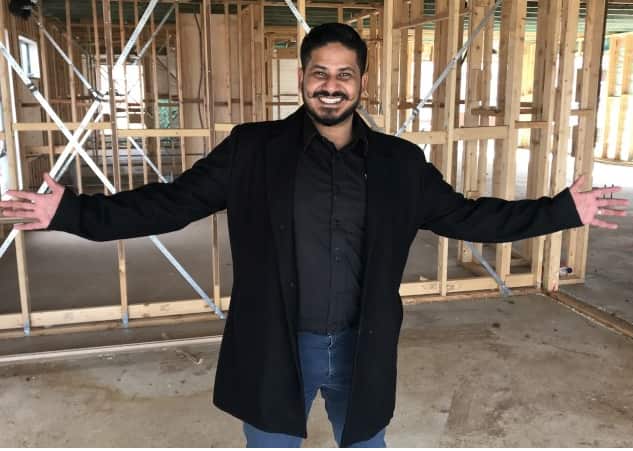 A smiling man with a handlebar moustache spreads his arms wide while standing in front of the frame of an unfinished house