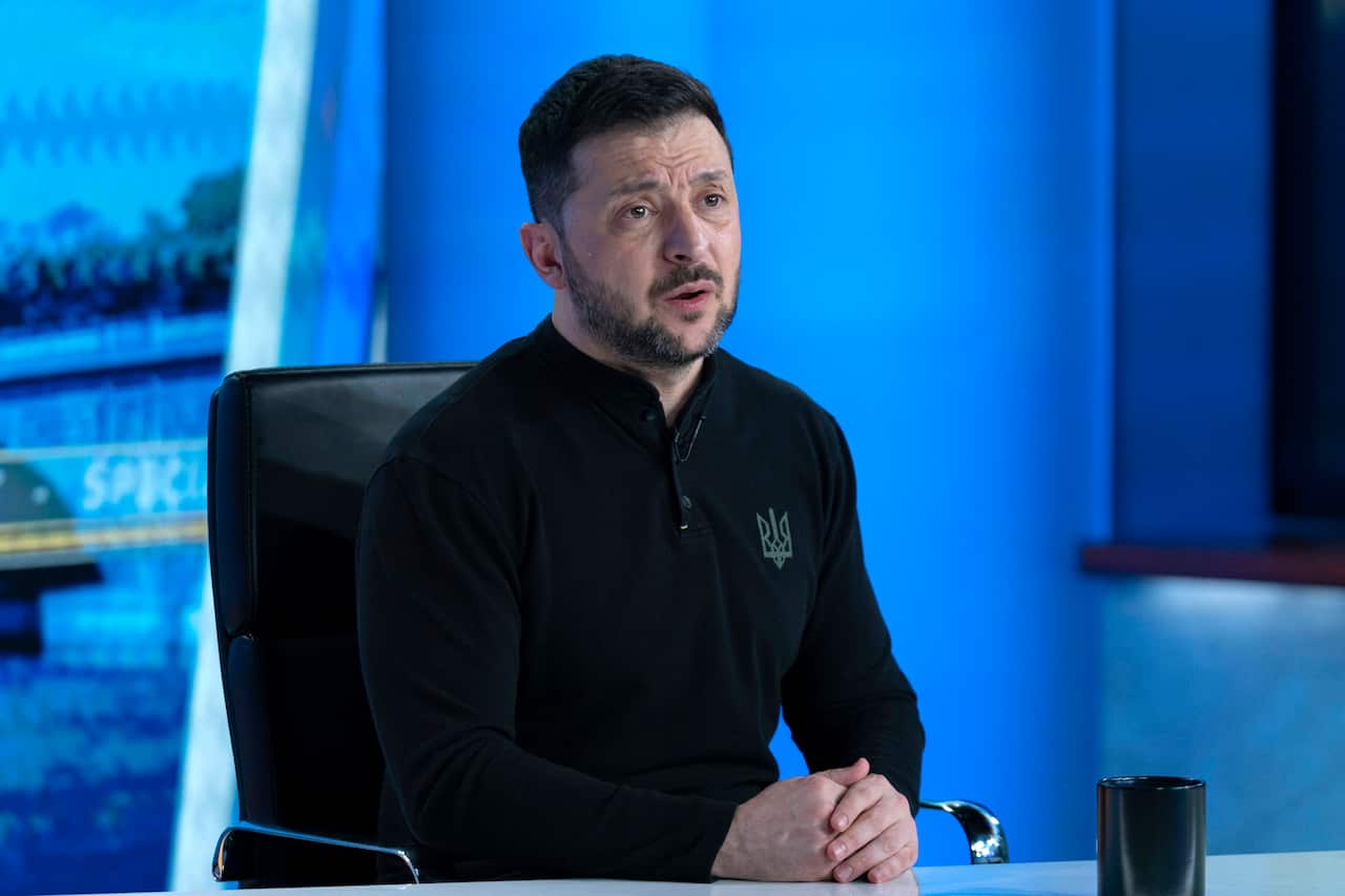 Ukraine's President Volodymyr Zelenskyy speaking during a TV interview. He's wearing a black long-sleeve top, sitting on a black chair and the set behind him is blue