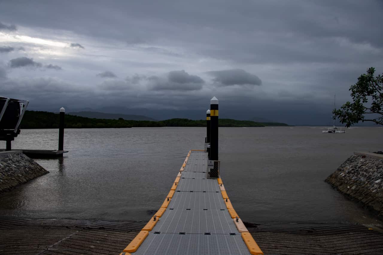 A floating grey and yellow boat dock extends into a calm bay under a heavy, overcast sky, flanked by stone embankments and distant green hills.
