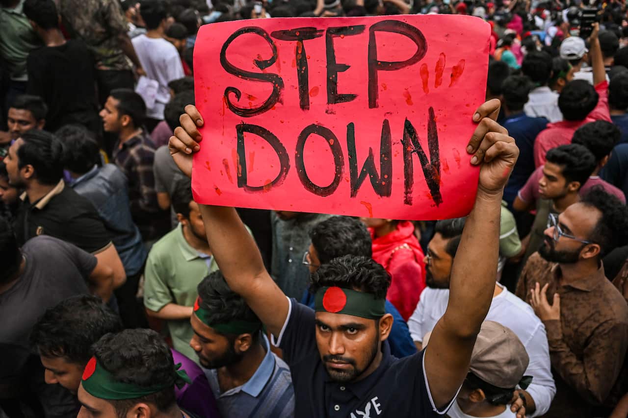 A young Bangladeshi man wearing a black headband with a red circle in the middle holds up a red cardboard reading "STEP DOWN". He is surrounded by many other young people, some of whom are wearing similar headbands. 