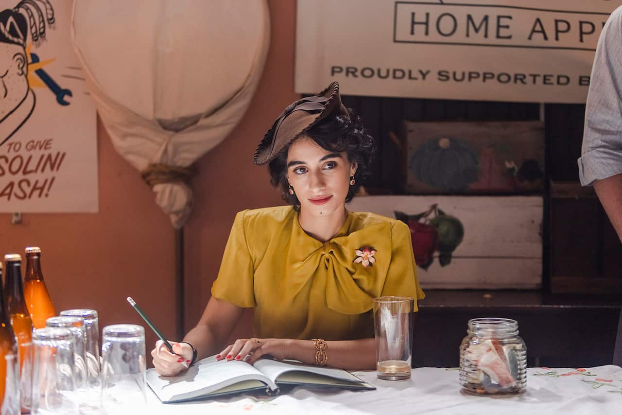 A woman in a yellow top and a jauntily angled brown hat sits at a desk, with a pencil in hand and a book in front of her. 