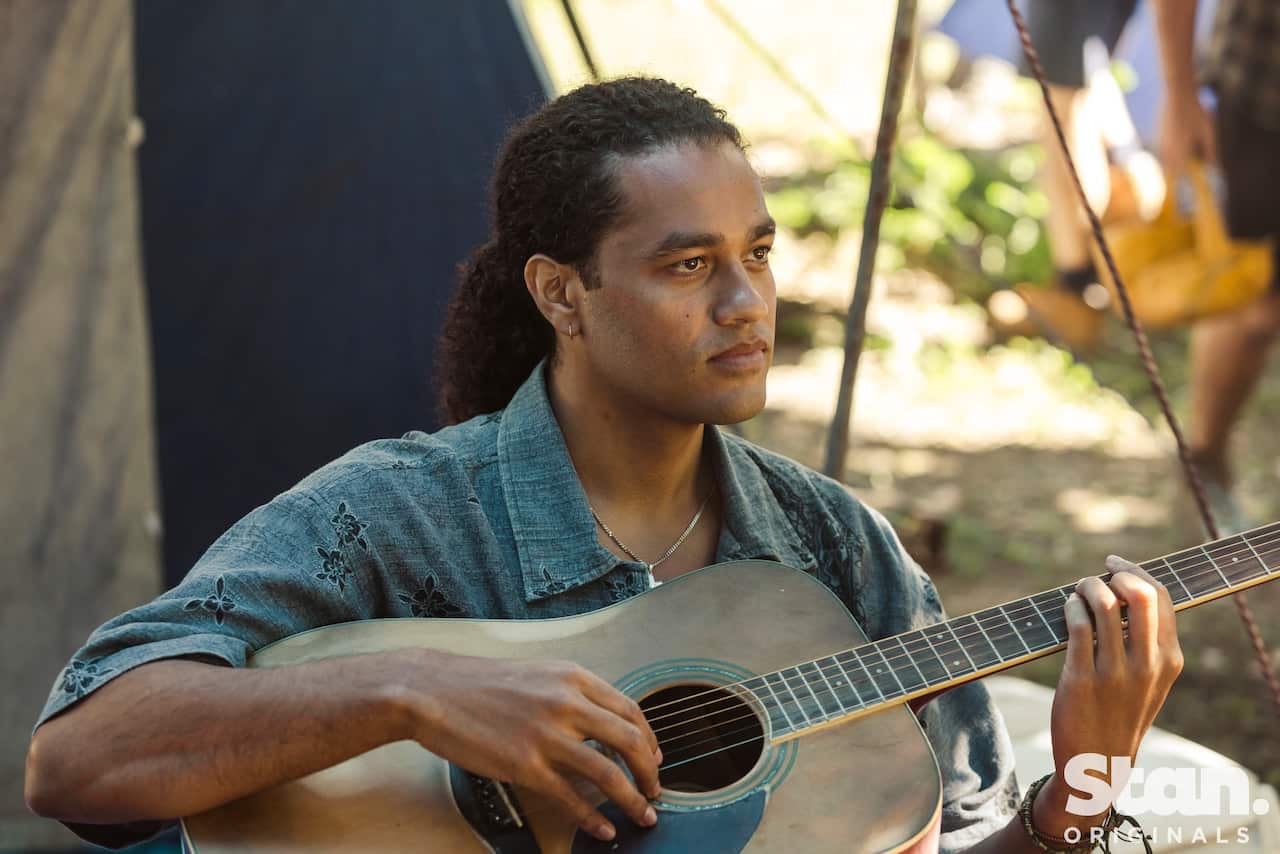 Ziggy Ramo sitting outside at a makeshift camp wearing a light grey island shirt and holding an acoustic guitar
