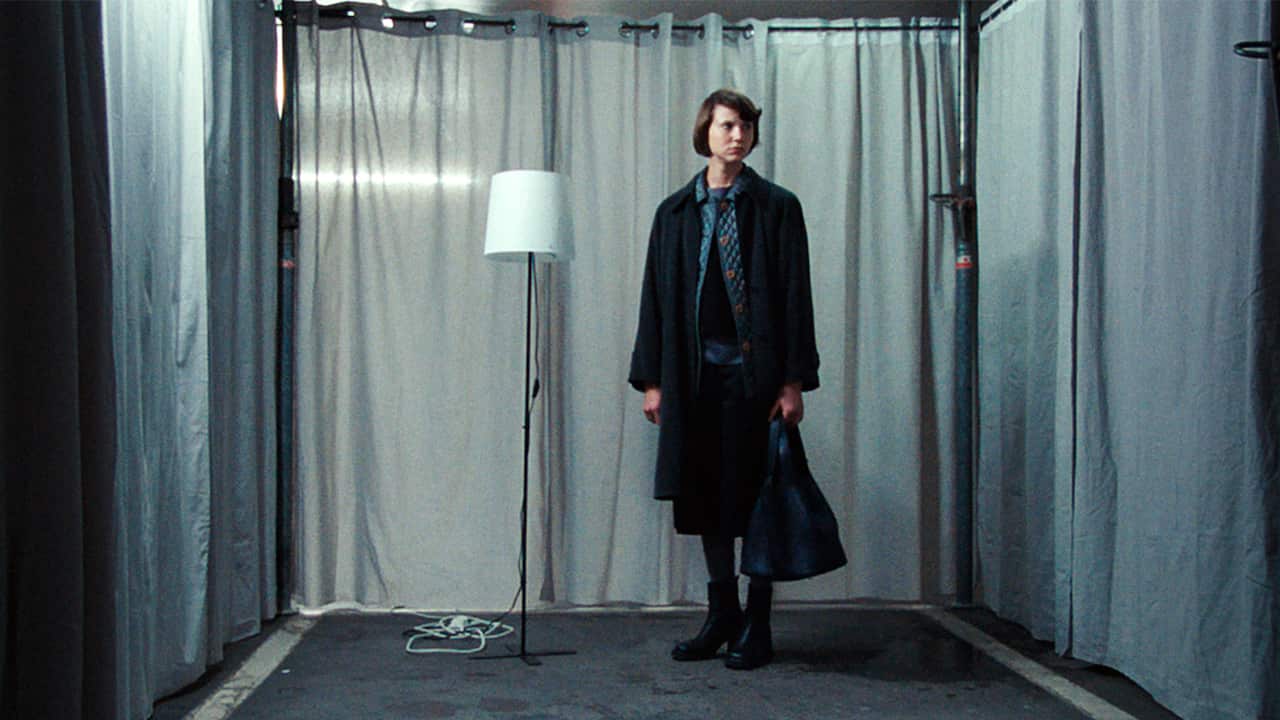 A woman in dark clothing stands in a small cubicle, with floor-length curtains forming the walls behind and beside her. The only other object in the cubicle is a tall lamp.  