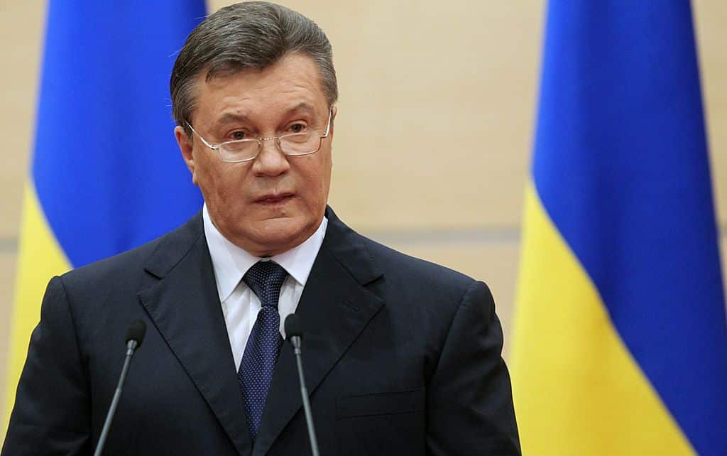 A man in a dark suit talking from behind two microphones. There are two Ukrainian flags behind him
