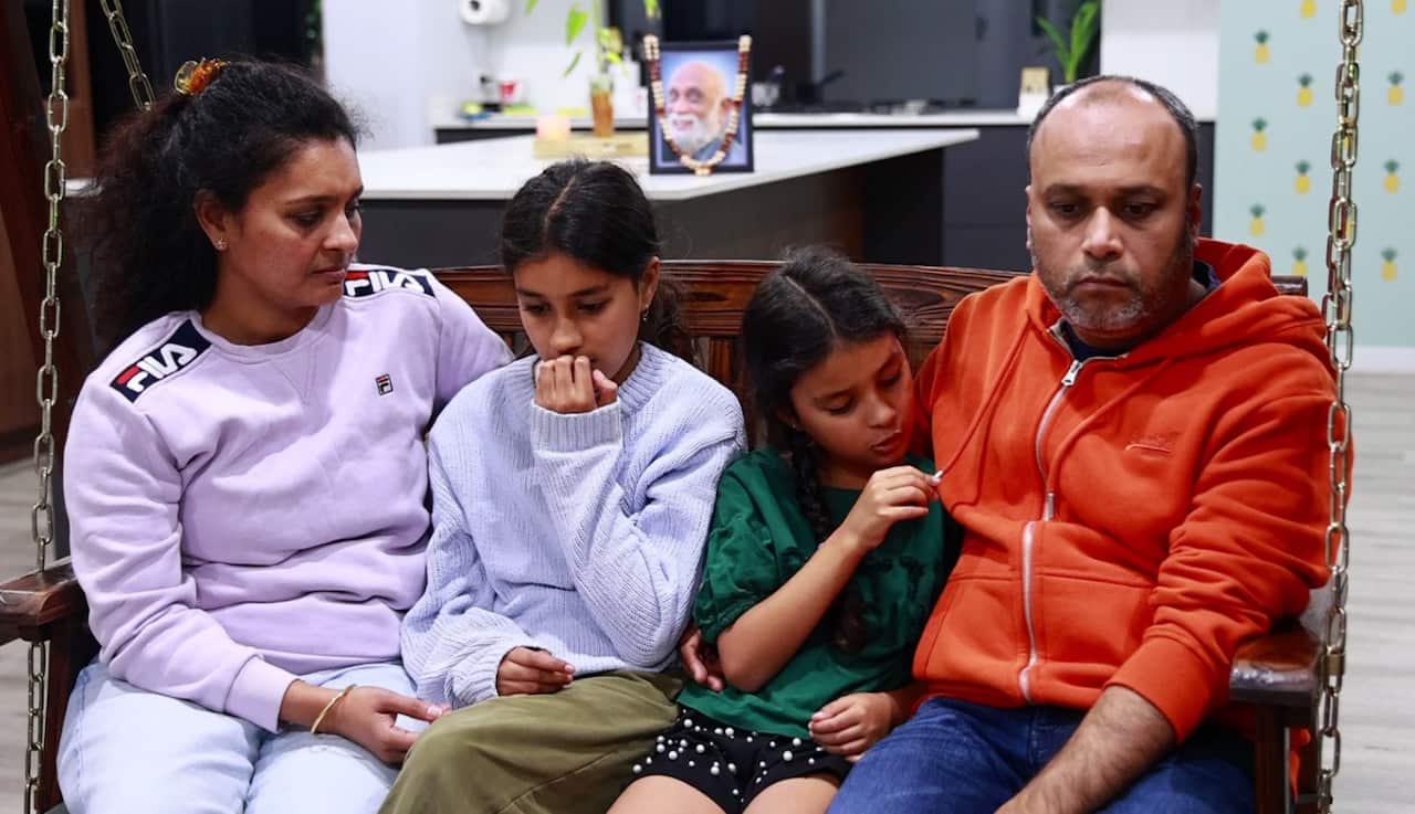 A man and a woman sit together on a swing in their family room, with their two daughters seated in between them. All four wear sad expressions.