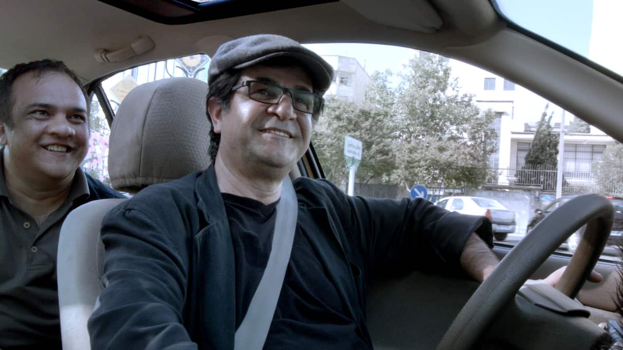 Two men smiling inside a car, with the driver wearing glasses and a flat cap.