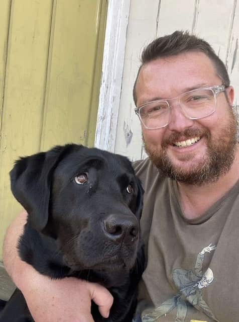 A man with beard and glasses hugs a black Labrador 