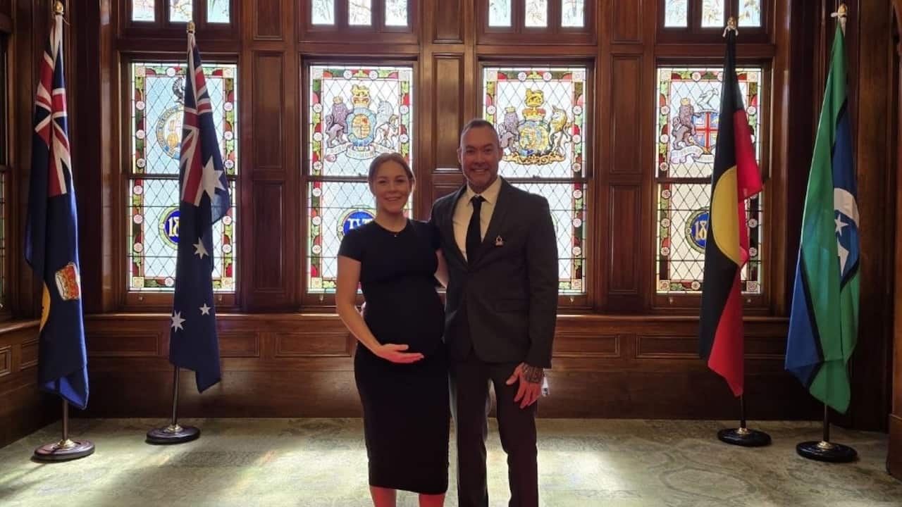 a middle aged couple dress in black formal attire in a government building with the Australian flag, Aboriginal flag, Torres Strait Islander flag and stained glass windows