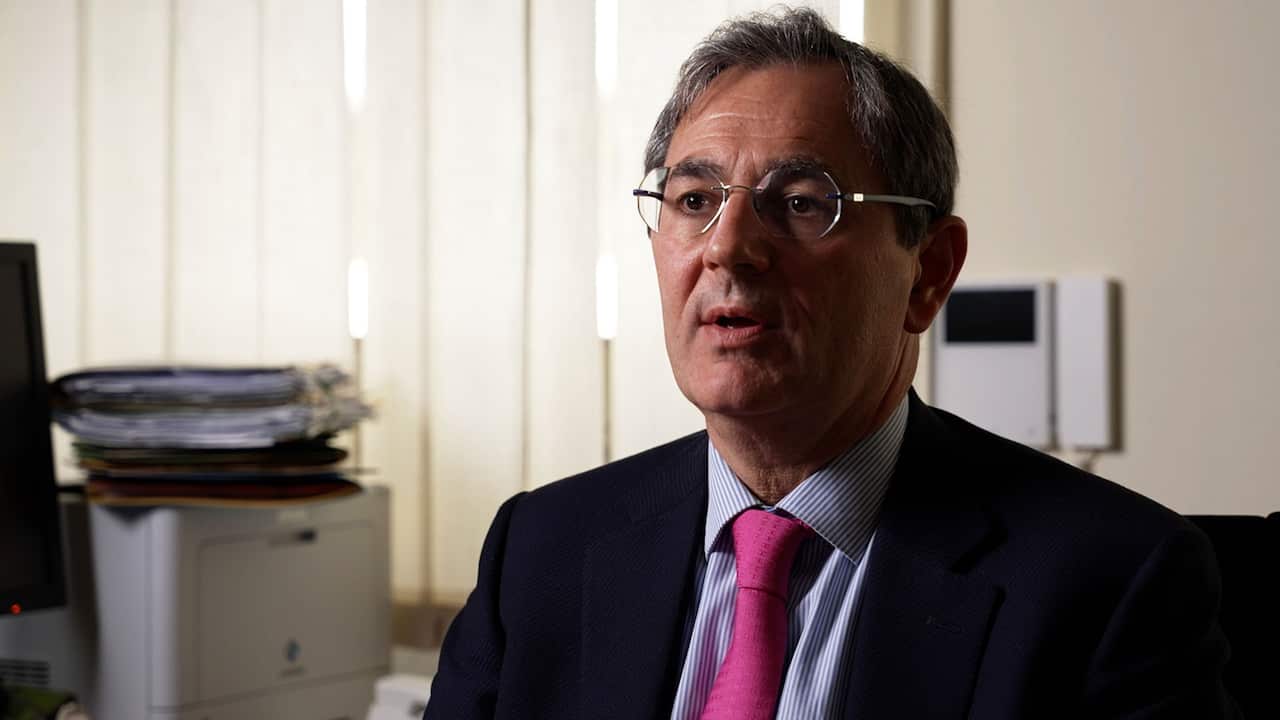 An older man with grey hair and reading glasses wearing a dark suit, blue shirt and a pink tie
